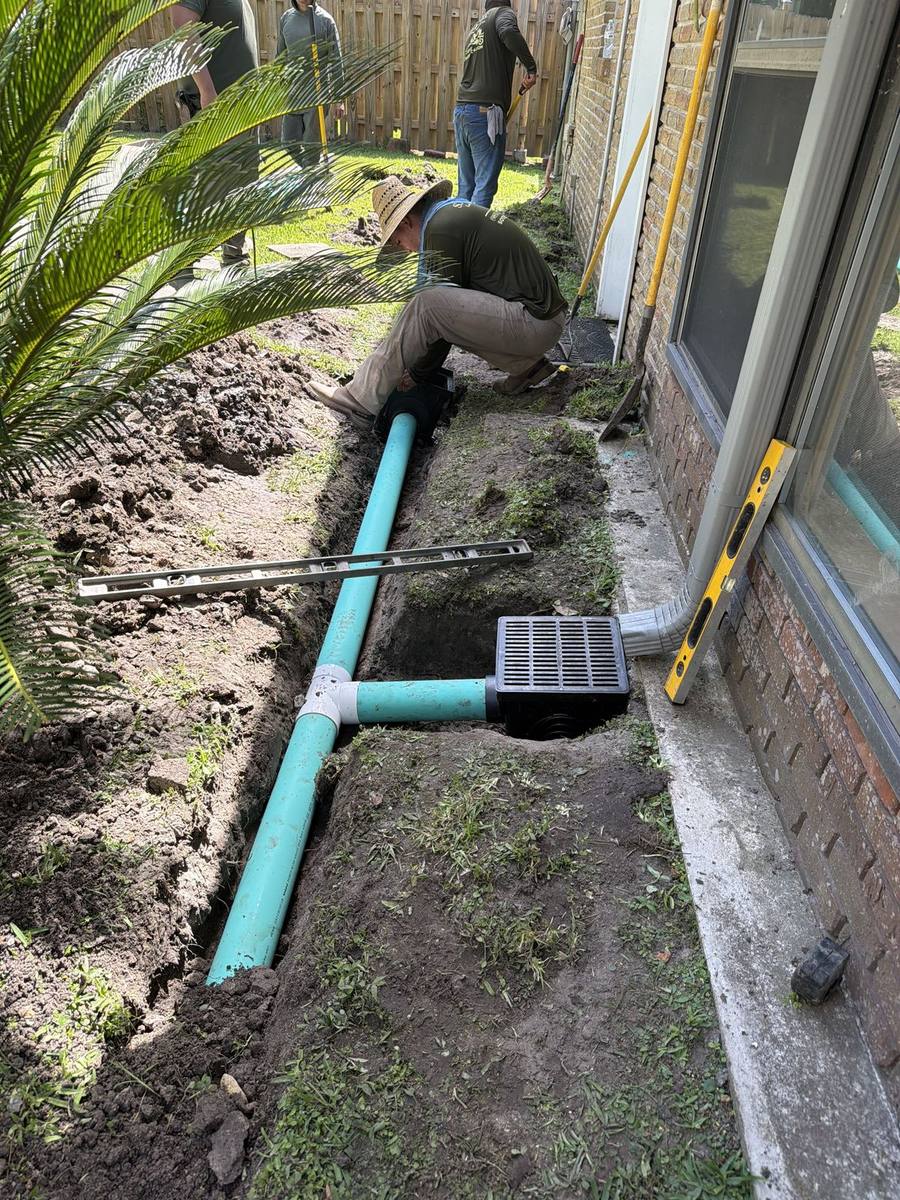 Drain line and catch basin installed along a patio edge to carry roof runoff away from the slab